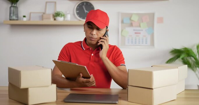 Portrait of Young Asian busy postman in red uniform and cap sitting at desk and talking on mobile phone in postal office store and looking at clipboard. Parcels front side.