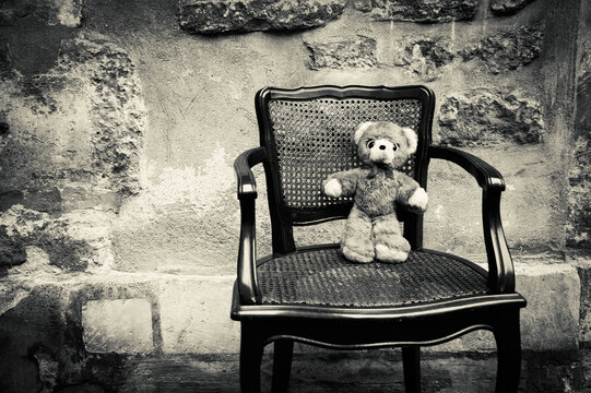 Teddy Bear sitting on chair and rough stone wall at background. Child care, abuse, children rights concepts. Black white historic photo