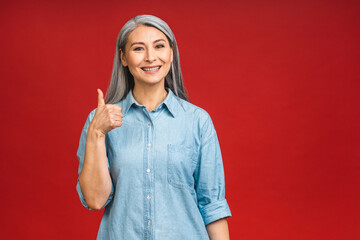 Happy elegant mature senior aged asian business woman smiling standing isolated over red background. Smiling confident cheerful middle aged lady, copy space. Thumbs up sign.