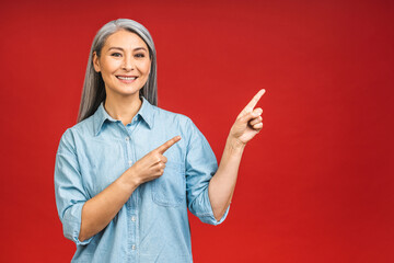 Portrait of attractive cheerful beautiful senior mature aged woman demonstrating copy space isolated over red background, pointing finger away.