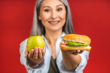 Close-up portrait of mature senior asian woman holding burger and green apple. Standing isolated over red background. Diet concept.