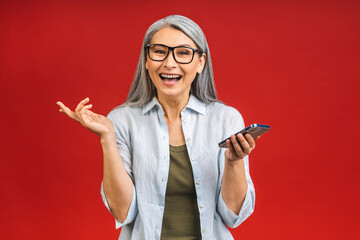 Photo portrait of asian shocked amazed surprised senior aged mature woman holding mobile phone seeing crazy discounts wearing casual isolated on red background.