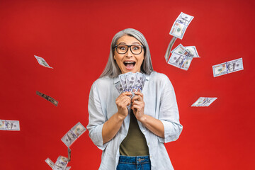 Happy winner! Image of a mature senior asian happy old woman standing isolated over red background, holding money.