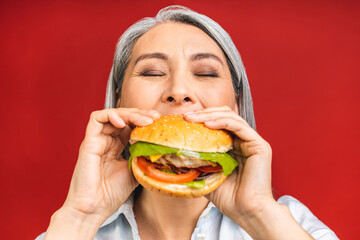 Mature senior woman eating burger with satisfaction. Grandmother enjoys tasty hamburger takeaway,...