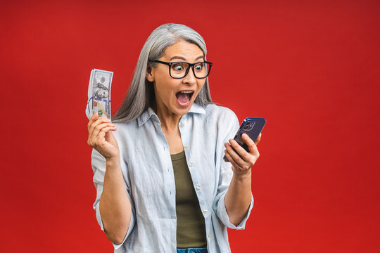 Happy winner! Image of mature aged happy asian senior woman standing isolated over red background wall  holding money using mobile phone.