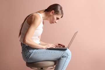 Woman with bad posture using laptop while sitting on stool against pale pink background - obrazy, fototapety, plakaty