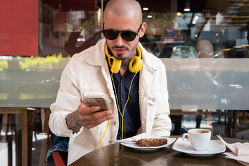 Young man with glasses and yellow headphones chats on his cell phone in a cafe drinking coffee and eating a cake