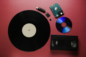 The evolution of recording and storing music and data, vinyl record, cassette, disk, hard drive, flash cards on a red-burgundy background, close-up, top view