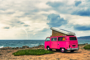 pink clasic camper van standing in front of the sea on a cliff with dramatic sky - travel concept.