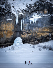 people ice fishing in front of an impressive rock wall with icicles hanging from it and forming an ice tower below.