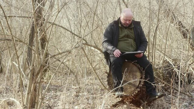 a man in a forest without leaves sitting on a large stump looks at his photos in his laptop-transformer