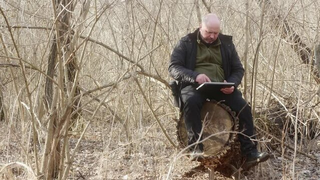 a man in a forest without leaves sitting on a large stump looks at his photos in his laptop-transformer
