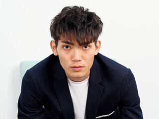 Portrait of handsome Chinese young man in dark blue leisure suit sitting in armchair and posing against white wall background. Looking at camera seriously, front view.