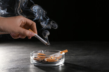 Woman holding smoldering cigarette over glass ashtray at grey table against black background,...