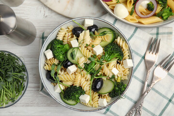 Bowl of delicious pasta with cucumber, olives, broccoli and cheese on white wooden table, flat lay