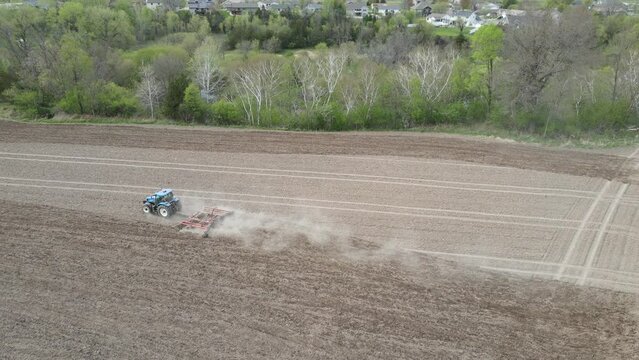 Drone view of farmer discing field in preparation for planting. Trees lining field with creek.  Pattern of lines through the field. Residential housing seen in distance. 