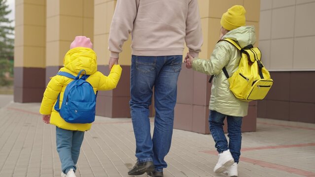 Dad Leads The Girls' Daughters By The Hand To School, Backpacks With Books On Shoulders Of Schoolgirls, Hold Older Father By The Hand, Pick Up Little Kid From School, Education And Upbringing Lessons