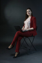 Fototapeta na wymiar A girl on a black background, in a red pantsuit and high heels, is sitting on a chair with a laptop on her lap. Programmer, freelancer, successful businesswoman or secretary