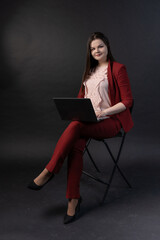 A girl on a black background, in a red pantsuit and high heels, is sitting on a chair with a laptop on her lap. Programmer, freelancer, successful businesswoman or secretary