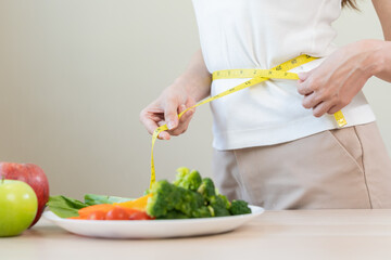 Slim shape asian young woman, girl standing, hand in measure tape around waist, body her have a vegetables on table as white background. Diet session for wellbeing health, eat good food people.