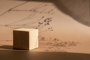 Geometric podium for demonstration of cosmetic products. Beige square 3d presentation stand on brown background in sunlight, with shadow from grass twigs