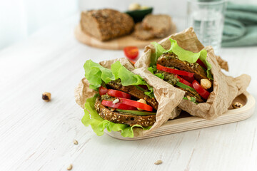 Making a snack to go Dietary vegan sandwiches with rye bread, peppers, tomatoes and vegetables wrapped in paper on a white wooden table. Ingredients in the background. Healthy eating. Space for text.