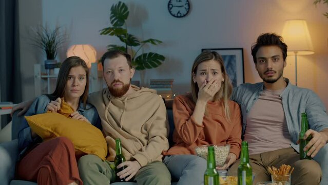 Shocked people diverse group watching thriller on TV eating popcorn relaxing in dark house together. Emotion and mass media concept.