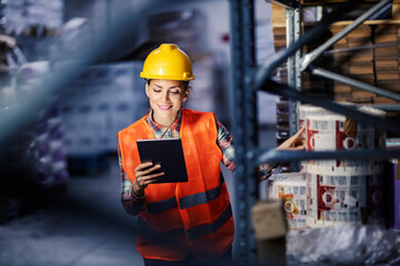 A happy female warehouse worker checking in inventory and smiling at tablet.