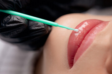 Master applies gel on red of lips of young woman after permanent makeup tattoo in beautician salon,...