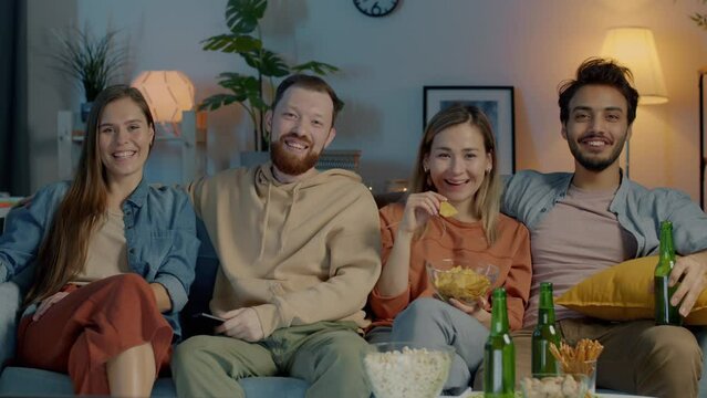 Portrait of diverse group of friends watching TV eating chips and laughing having fun together in dark house. Friendship and entertainment concept.