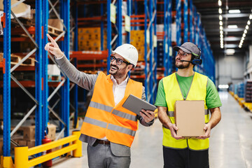 A supervisor pointing while warehouse warehouse worker with box looking in that way.