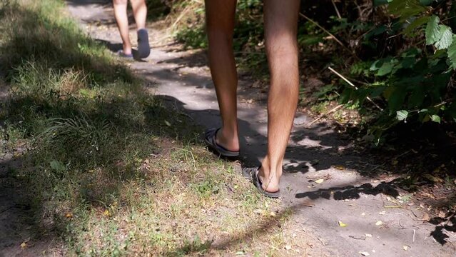 Dad and Son are Walking Along a Sandy Path in a Green Woods. Bare male and children feet in flip flop slowly step forward. Summertime. Shade from trees in the sunlight. Walk in fresh air. Relaxation.