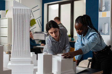 Architectural engineers doing teamwork reading construction plans over table with white foam scale model of buildings. Team of two architects colleagues collaborating looking down at blueprints.