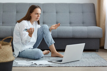 Image of confused helpless woman wearing white shirt and jeans sitting on floor near gray sofa,...