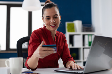 Employee in red shirt cheking credit card details for online shopping. Businesswoman at startup desk using using laptop for internet banking. Entrepreneur typing account number sitting at the office.