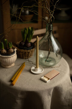 Tall Yellow Candle In Ceramic Candlesticks On The Table. A Tall Candle On A Table With Houseplants And A Large Transparent Glass Vase.