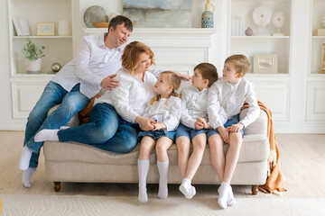 Big happy family sitting on couch in bright living room. Family portrait parents and children. Caucasian parents communicate with their daughter and two sons at home dressed in white shirts and jeans