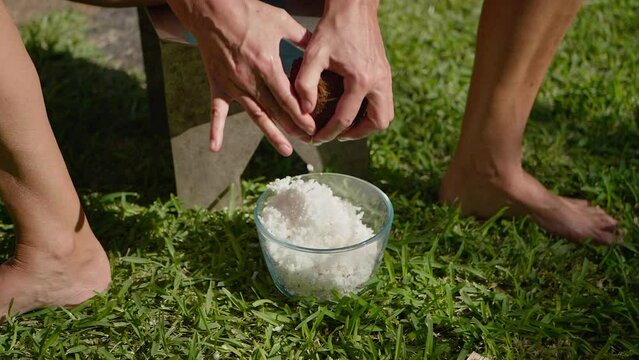 Slow motion close up of woman shredding fresh coconut into bowl using hand scraper.