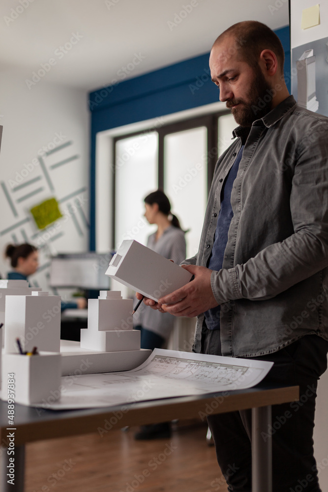 Poster building architect holding foam machette in hands looking for inspiration standing next to desk with