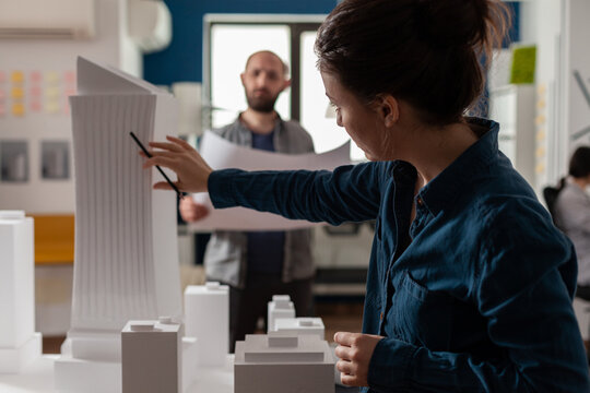 Architect inspecting futuristic building design of skyscraper with colleague engineer holding blueprints. Architects doing teamwork at desk with white foam maquette of construction project. - Powered by Adobe