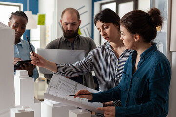 Two women architectural engineers looking down at blueprints next to colleagues holding laptop and tablet computer. Group of focused professional architects analyzing design plans.