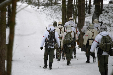 Soldiers in winter white camouflage coats are walking along a forest road. The German Army during World War II. Military historical reconstruction in winter.
