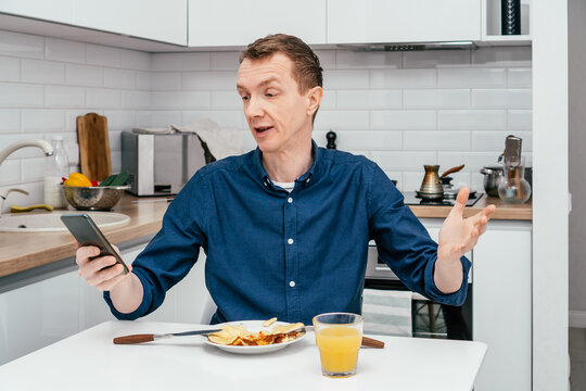 Portrait of surprised, astonished middle-aged man with short ginger hair in blue shirt looking at mobile phone with arm raised in front of omelette on plate near glass of orange juice in kitchen.