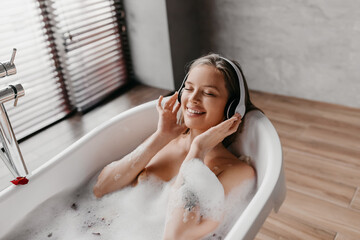 Charming woman lying in hot bubbly bath and listening to music in wireless headphones, enjoying...