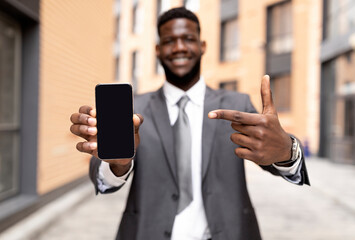 Business ad concept. African american businessman in formal suit pointing at smartphone with blank screen, mockup