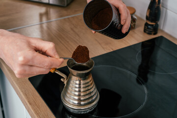 Close up of hands of unrecognizable man, putting full heaped tea spoon of natural ground coffee from round glossy tin in copper cezve on black electric cooker for brewing near spice grinder in kitchen