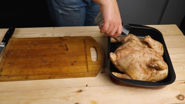 A woman cook pours water from a glass into a baking tray with spiced chicken. Cooking at home
