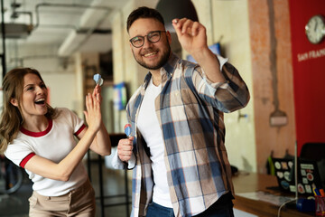 Young colleagues taking break after work. Happy young businessman and businesswoman playing darts...
