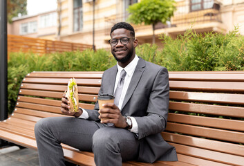 Enjoy your lunch. Happy african american businessman drinking coffee and eating sandwich, sitting on bench