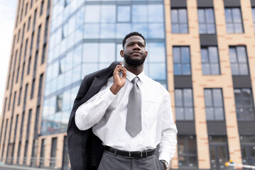 Young african american office worker holding jacket on shoulder and walking near modern office building, looking away